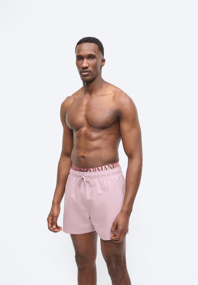 Μυώδης άνδρας όρθιος, φοράει ανοιχτό ροζ μαγιό με σχεδιασμένη ζώνη, σε λευκό απλό φόντο, με τα χέρια χαλαρά δίπλα στο σώμα.