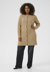 Cardigan long en tricot beige avec une texture douce, doté d'une fermeture à boutons sur le devant et de manches longues, associé à un pantalon noir à jambes larges.