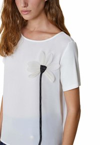 Top corto bianco a maniche corte realizzato in un tessuto trasparente, con una grande applicazione di fiore bianco e un accento in raso nero per il gambo. Design semplice ed elegante.