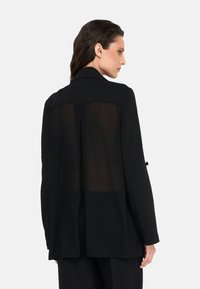 Blazer nero con pannello posteriore trasparente, maniche lunghe, colletto e dettagli con bottoni. Tessuto liscio e vestibilità sagomata.