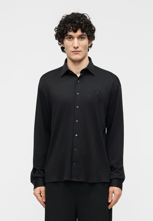 Homme aux cheveux bouclés portant une chemise noire à manches longues avec un blason brodé discret sur la poitrine gauche, se tenant devant un fond uni.