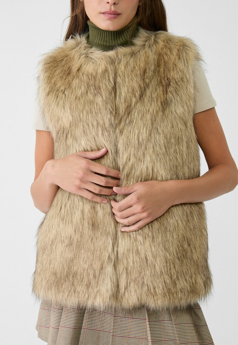 Gilet en fausse fourrure beige avec une texture douce. Couture verticale sur le devant, design sans manches, superposé sur un col roulé olive à côtes.