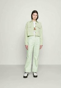 Mulher de pé contra uma parede lisa, vestindo um casaco curto verde claro, calças leves verdes claras com padrão ondulado, uma t-shirt gráfica branca e sapatos brancos chunky.