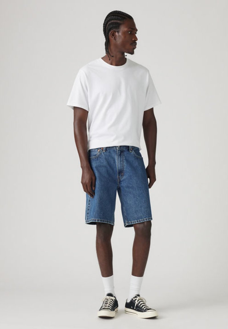 Homme aux cheveux tressés portant un t-shirt blanc, un short en jean bleu, des chaussettes blanches et des baskets noires, debout devant un fond uni.