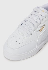 Hvite sneakers med skinn-overdel, perforert tåboks, flate hvite snørebånd og gulllogo detaljer på siden. Gummisåle for bra grep.
