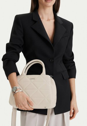 Kobieta w czarnej marynarce trzyma beżową pikowaną torebkę z podwójnymi uchwytami i nosi srebrny zegarek na nadgarstku na jednolitym tle.