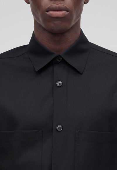 Gros plan d'une personne portant une chemise noire à boutons avec un col pointu et deux poches poitrine visibles sur un fond uni.