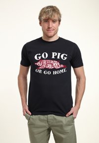 Svart t-shirt med grafiken "GO PIG OR GO HOME" i vit och röd text med matrelaterade ord, i kombination med ljusgröna byxor.