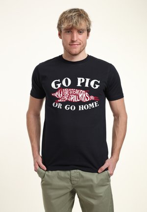 Czarny T-shirt z grafiką "GO PIG OR GO HOME" w białym i czerwonym kolorze z jedzeniem związanymi słowami, w połączeniu z jasnoszarymi spodniami.