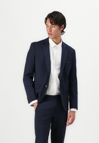 Jūrų mėlynos spalvos švarkas su notch apykakle, vienu mygtuku ir dviem priekinėmis kišenėmis, derinamas su balta marškinių chemise. Slim fit, modernus dizainas.
