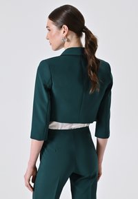 Blazer a manica tre quarti di colore verde smeraldo, con texture liscia e pannello bianco in satin in vita, abbinato a pantaloni coordinati.