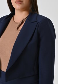Marineblazer met een getailleerde pasvorm, voorzien van een geknoopte kraag en schoudervullingen. Daaronder een beige top met een geknoopte halslijn.