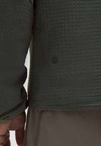 Donkergroene textured jas met een ruitpatroon, voorzien van een klein zwart logo aan de zijkant. De manchetten zijn elastisch en de details zijn minimaal.