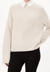Beige Pullover mit gerippter Textur, darunter ein weißes Hemd mit Kragen. Lockerer Schnitt mit langen Ärmeln, endend auf Hüfthöhe.