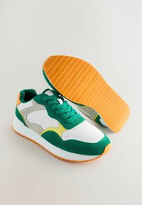 Adidași verzi și albi cu parte superioară din suede și plasă, accente galbene, talpă din cauciuc portocalie și croială cu șireturi.