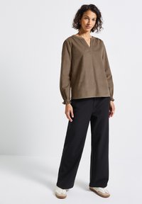 Donna in piedi che indossa una blusa marrone a maniche lunghe, pantaloni larghi neri e sneakers bianche su uno sfondo bianco semplice.