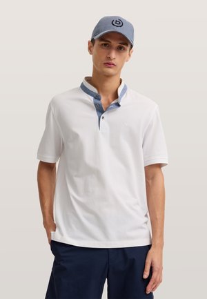 Giovane uomo che indossa una polo bianca a maniche corte con dettagli blu sul colletto, pantaloncini blu navy e un berretto grigio con logo nero su sfondo neutro.