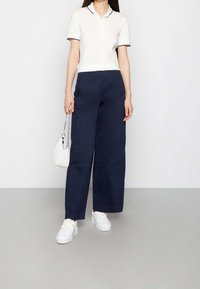 Vit polo skjorta med marinblå detaljer, i kombination med vida marinblå byxor. Modellen håller en vit vadderad handväska och bär vita sneakers.