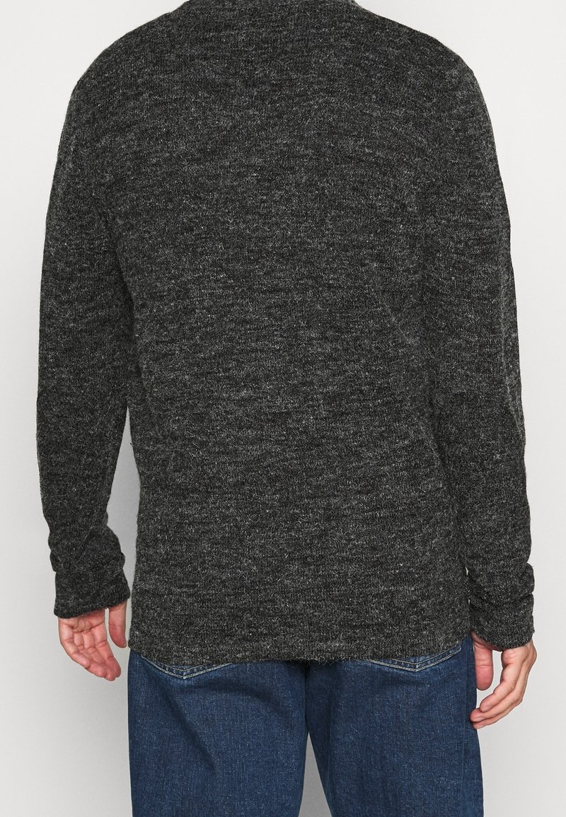 Person som bär en mörkgrå stickad tröja och blå jeans, visad bakifrån mot en enkel ljus bakgrund.