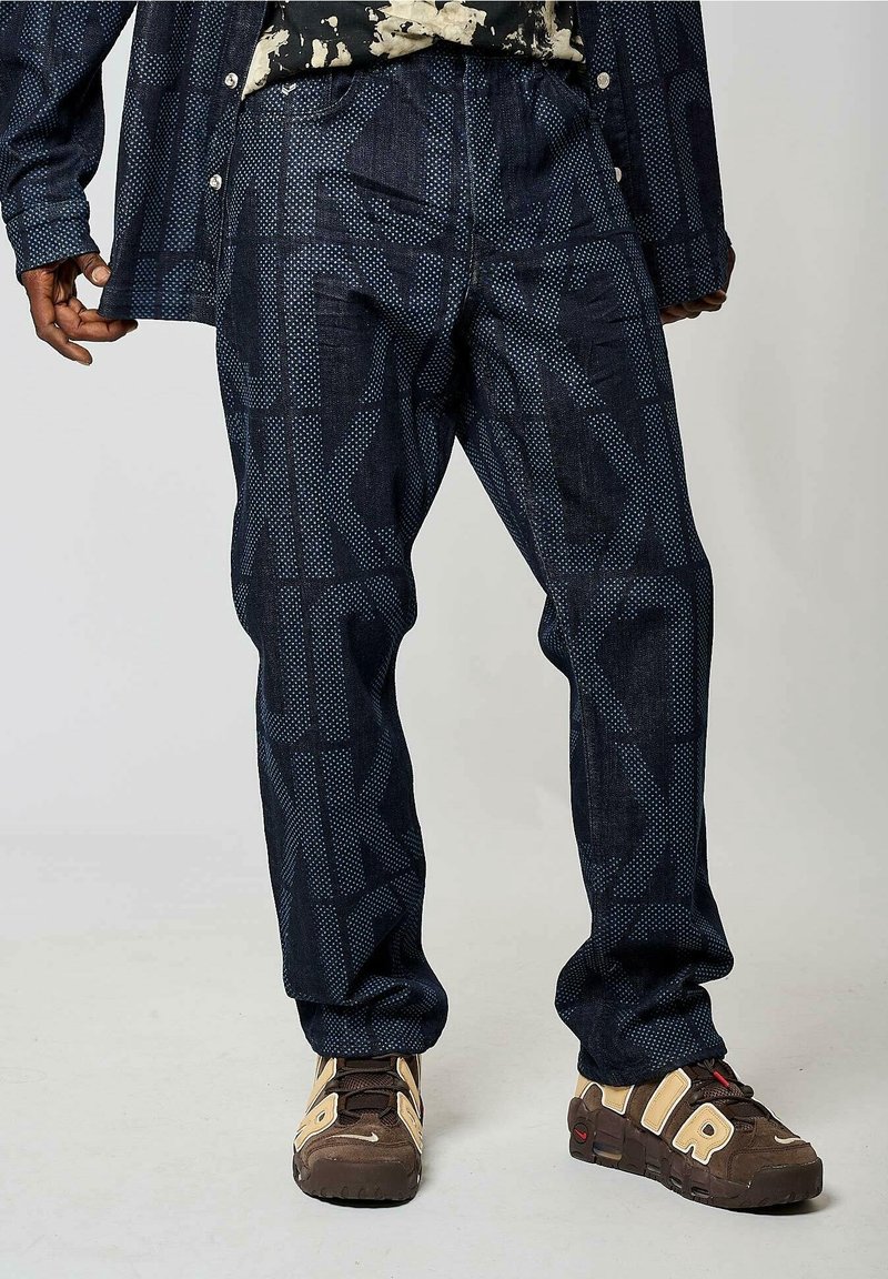 Personne portant un pantalon bleu foncé à motif, une chemise assortie, un haut à motifs, et des baskets marron avec des détails beige et rouge.