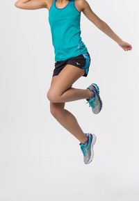 Un débardeur turquoise associé à un short noir. Des chaussures de course grises présentent des accents turquoise et violets. La personne est en plein saut, jambes levées.