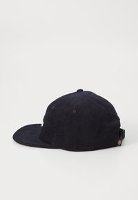 Gorra de pana azul marino con visera plana, construcción de seis paneles, botón superior y correa ajustable. Presenta un logo de marca lateral.