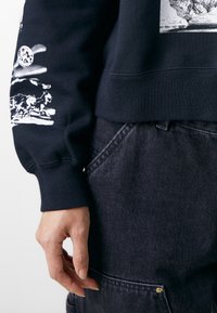 Felpa blu navy con grafiche bianche sulle maniche e in cotone misto. Abbinata a pantaloni di denim scuro con cuciture visibili e tasche.
