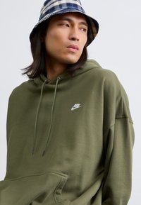 Ελιά πράσινη hoodie με μπροστινή τσέπη, κορδόνια και ένα μικρό λευκό λογότυπο. Το μοντέλο φοράει ένα καπέλο bucket με σχέδιο καρό σε ναυτικό μπλε και λευκό.