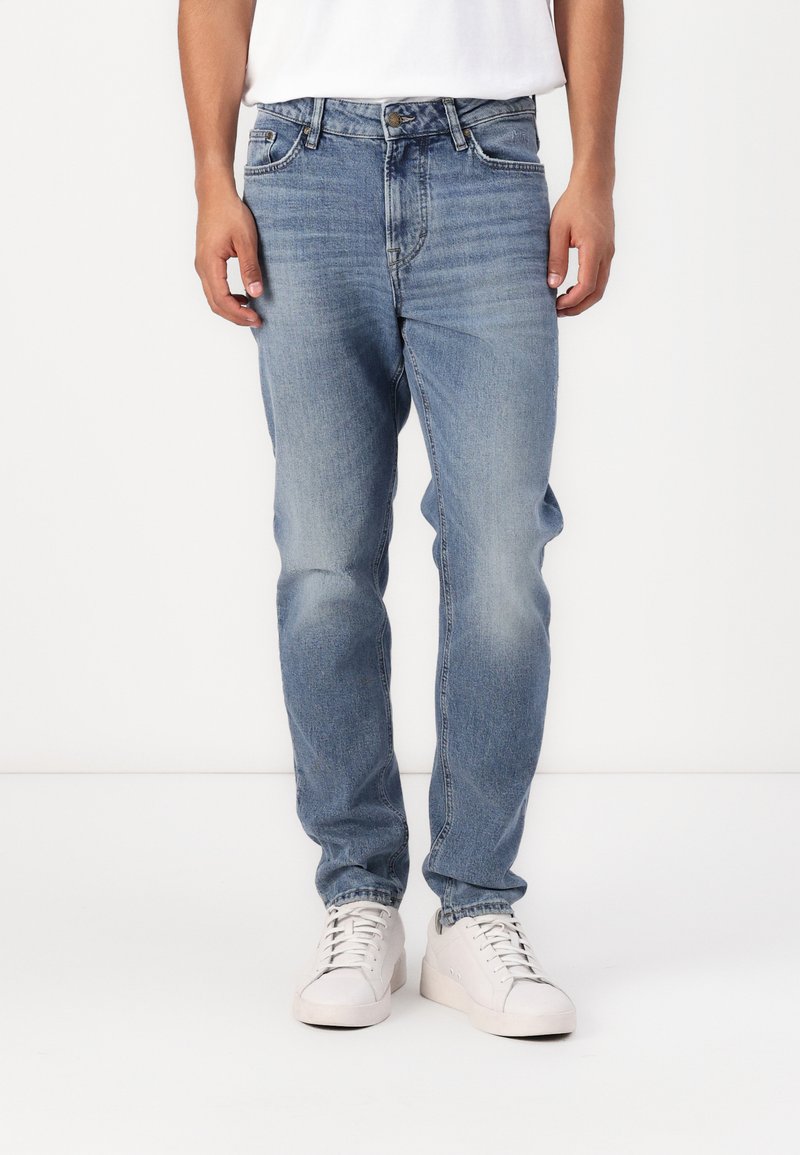 Jeanși din denim de culoare bleu deschis, cu o croială dreaptă, cinci buzunare și decolorare minimă. Purtați cu adidași albi, creând o apariție casuală.
