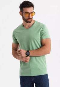 T-shirt verde a V in tessuto morbido, abbinato a jeans scuri. Il modello indossa occhiali da sole gialli e un orologio nero con cinturino testurizzato.