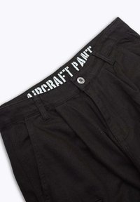 Pantalon noir en tissu texturé, avec une fermeture à bouton et une ceinture étiquetée « PANTALON AÉRIEN » en lettres blanches en gras.