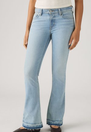 Jean évasé bleu clair avec des ourlets effilochés, porté avec des chaussures noires et un haut blanc sans manches, visible de la taille aux pieds.