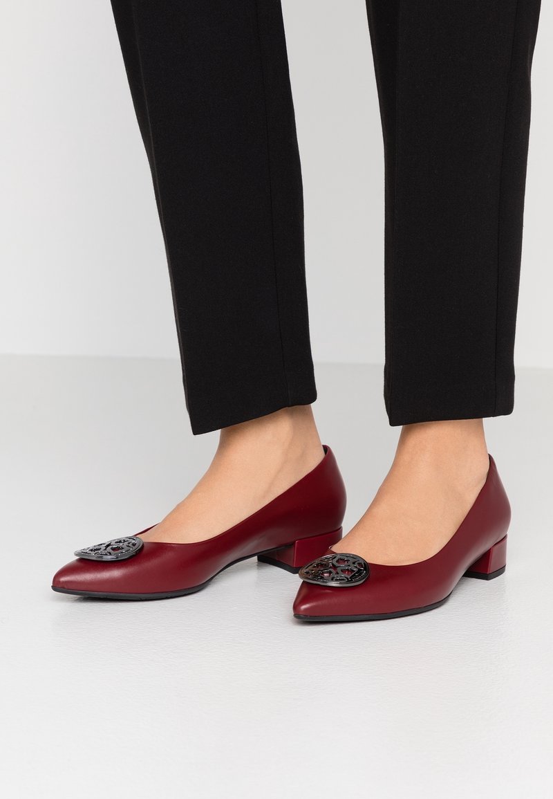 Femme portant des chaussures bordeaux à talons bas avec des médaillons décoratifs noirs associées à un pantalon noir ajusté sur un sol clair.