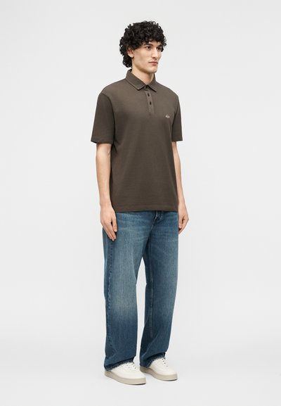 Jeune homme debout, vêtu d'un polo marron foncé, d'un jean bleu et de baskets blanches, sur un fond clair uni.