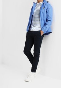 Jasnoniebieska kurtka przeciwdeszczowa z kapturem, biało-granatowa koszulka w paski, czarne spodnie slim fit i białe sneakersy. Kurtka z logo.