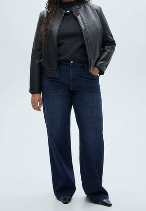 Femme portant une veste en cuir noire, un haut noir, un jean bleu foncé à jambes larges et des chaussures noires pointues, debout avec une main dans la poche.