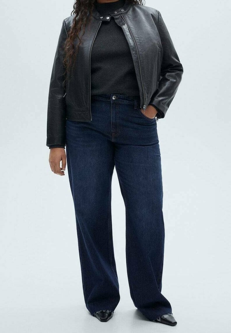 Femme portant une veste en cuir noire, un haut noir, un jean bleu foncé à jambes larges et des chaussures noires pointues, debout avec une main dans la poche.