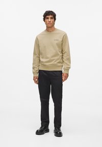 Béžová mikina s rebrovanými manžetami a spodným okrajom, s malým čiernym logom na hrudníku, v kombinácii s čiernymi nohavicami a čiernymi topánkami.