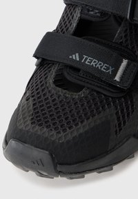 Sort sportssko med mesh-overdel og velcro-strop, der har en tekstureret sål og logo-detaljer. Nærbillede fremhæver materiale og design.