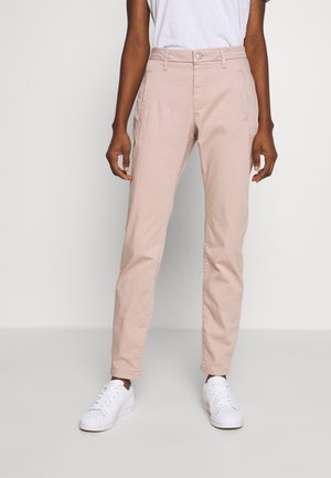 Persoon die lichtroze broek met rechte pijpen en witte sneakers draagt, staand tegen een effen lichte achtergrond.