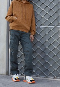Persona con capucha marrón de estampado, pantalones cargo verde oscuro y zapatillas blancas con suelas naranjas, de pie junto a una puerta de metal gris con patrón.