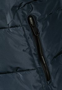 Musta vetskari vetotapilla tummansinisiä tikattuja kangasmateriaaleja vasten, korostaen takin taskun yksityiskohtia ja ompelua.