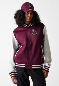 Burgunder varsityjakke med grå ermer, stripete ribbede mansjetter, hvite detaljer og LA-logo. Kombinert med en matchende burgunder caps med blå logo.