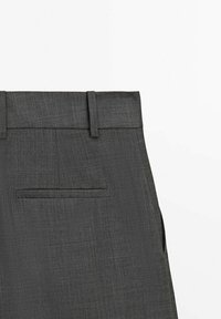 Pantalon gris foncé en tissu texturé, avec une taille plate, des passants de ceinture et une poche arrière unique. Design ajusté.