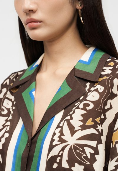 Γυναίκα που φορά καφέ πουκάμισο με πράσινα, μπλε και λευκά γεωμετρικά και αφηρημένα σχέδια και χρυσούς κρίκους στα αυτιά, εμφανιζόμενη από τον λαιμό έως τους ώμους.