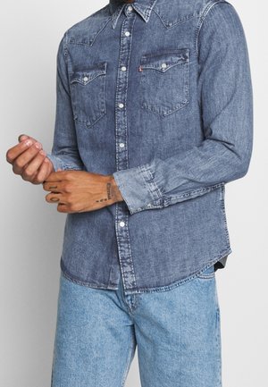 Mann richtet den Ärmel eines ausgewaschenen blauen Denim-Knopfhemds mit Druckknopftaschen zurecht, kombiniert mit hellblauen Jeans vor einem schlichten Hintergrund.