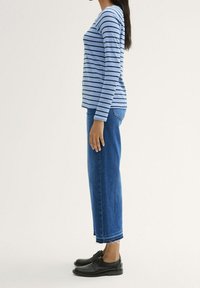 Modrá a bílá pruhovaná košile s dlouhým rukávem v kombinaci s vysokým pasem modrých džínů a černými botami, zobrazeno z boku na neutrálním pozadí.