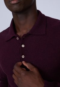 Polo en tricot bordeaux avec col, patte de boutonnage à trois boutons et tissu texturé. Gros plan avec une main sur la chemise.