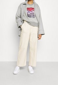 Šedá oversized bunda, grafická mikina s designem hor, krémové široké kalhoty, bílé tenisky. Hladké textury, uvolněný střih.