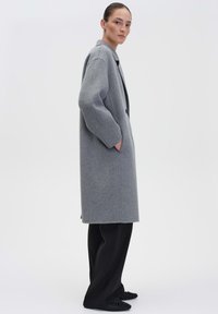 Grijze wollen overjas met een relaxte pasvorm, grote kraag en zijzakken. Gecombineerd met zwarte broek en platte schoenen. Soepele textuur.